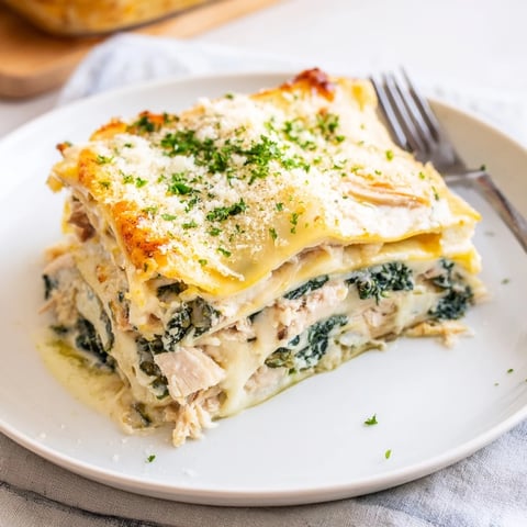 A close-up of golden, bubbly Chicken Alfredo Lasagna slice on a white plate, garnished with fresh parsley.